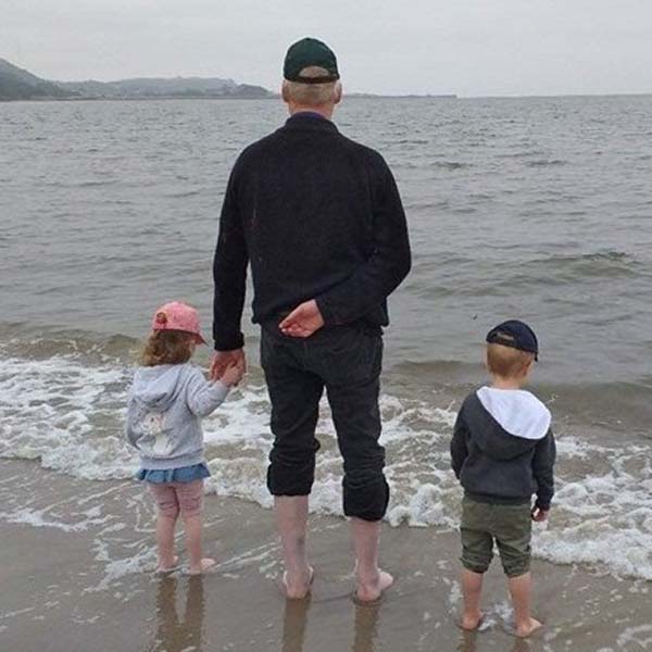A man and two young children holding hands whilst paddling near the shore at the seaside.