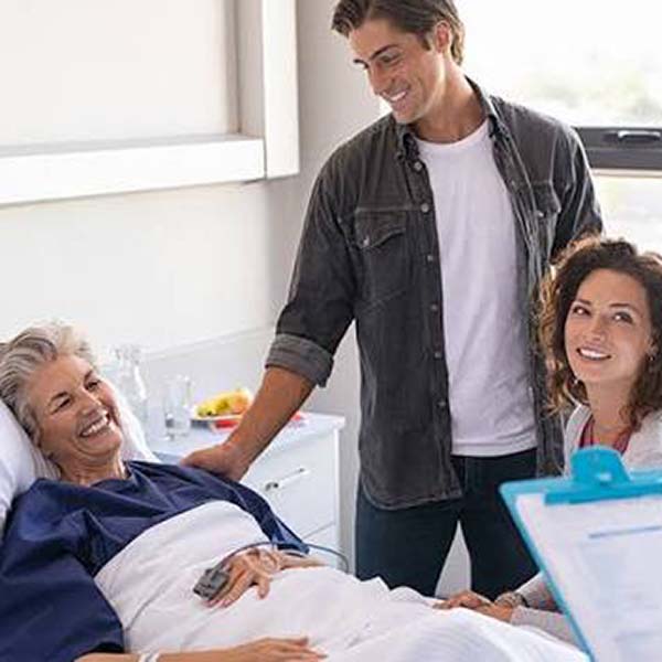 An elderly female patient sitting up in a medical bed with two relatives visiting her in the room.